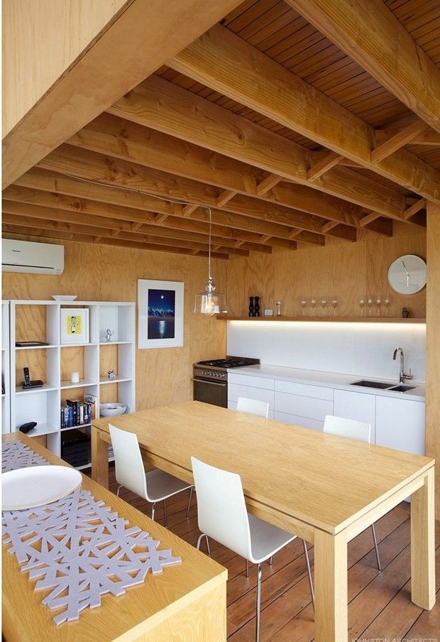 A compact open-plan kitchen and dining area with extensive warm plywood wall and ceiling finishes, exposed timber roof framing, and a large light-wood dining table surrounded by white chairs. The minimalist white kitchen includes flat-panel cabinetry, a dark cooktop and oven, an undermount sink with chrome tap, and a softly illuminated backsplash shelf. Open shelving, pendant lighting, framed artwork, and a wall clock complete the space.