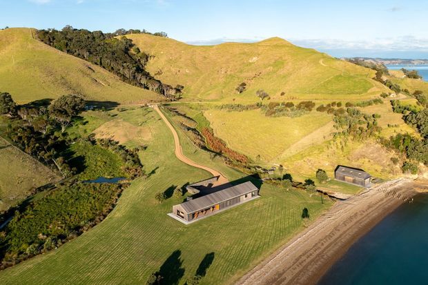 Aerial view of a long, low modern building set within expansive rolling green farmland beside a sandy shoreline and calm blue water. The property includes a second smaller outbuilding, landscaped lawns, a pond or wetland area, internal dirt tracks, and surrounding wooded hills. The main building has a simple dark pitched roof, extensive glazing, and a linear form integrated into the rural coastal landscape.