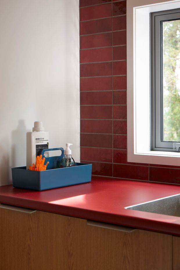 Close-up of a compact kitchen or utility workspace featuring a vivid red solid-looking benchtop, matching glossy red rectangular tiled splashback, warm wood-grain lower cabinetry, and a dark-framed window with white trim. A stainless-steel sink is partially visible at the right, while a blue storage tray with cleaning supplies sits on the counter. Natural daylight enters through the window.
