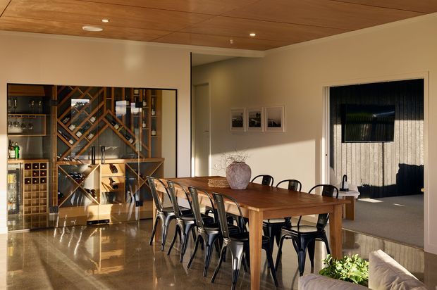 A warm, refined dining area with a large timber dining table and black metal chairs, opening into an adjacent living room. A substantial built-in wine display and storage unit occupies the left wall, while polished stone flooring, a timber-lined ceiling, recessed lighting, and neutral plaster walls create a contemporary entertaining space.