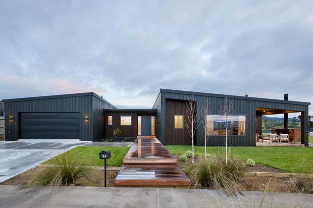 Single-storey contemporary residential home viewed from the street, with a dark vertically ribbed exterior, low-pitched rooflines, an integrated double garage, and a central covered entry. A timber walkway crosses a landscaped front garden, while large windows and an open covered outdoor area reveal a dining setting and distant rolling landscape.