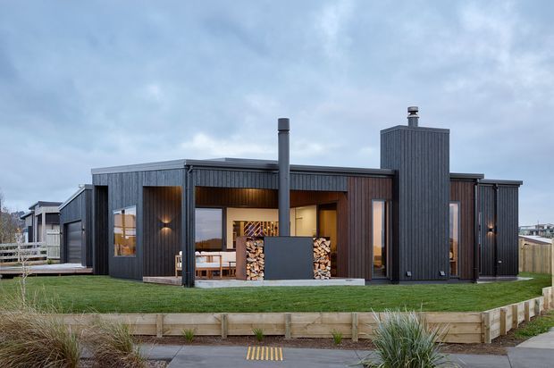 Single-storey contemporary dwelling viewed from the front, with dark vertically ribbed cladding, a flat roof, projecting chimney, extensive glazing, and a recessed sheltered entry or outdoor living area. Timber retaining edging, lawn, and coastal-style grasses frame the foreground.