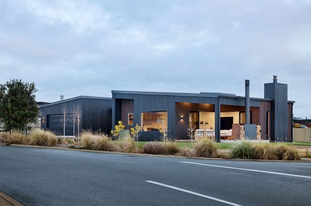 A low, contemporary single-storey house viewed from across a paved road. The facade combines dark vertical corrugated cladding with warm timber-lined recessed openings and large glazed living areas. A simple flat or very low-pitched roofline, tall chimney elements, and native-style ornamental grasses and shrubs form the front landscape under an overcast sky.