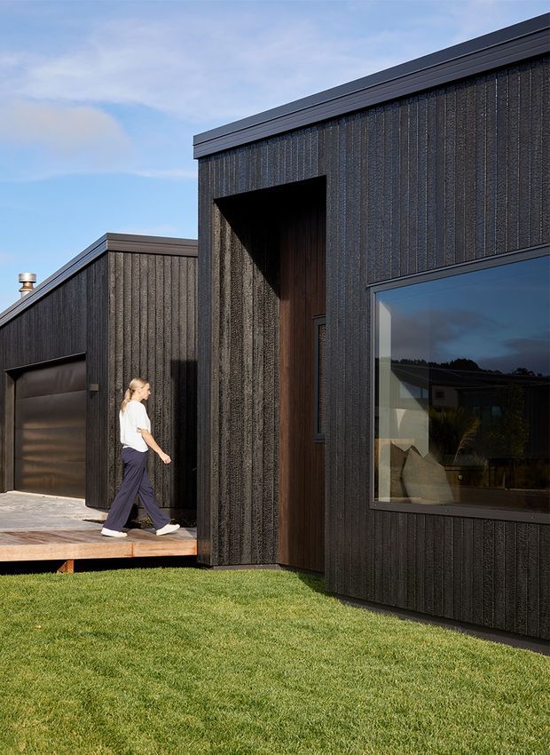 Exterior view of a contemporary single-storey home with dark vertically textured cladding, crisp flat rooflines, a recessed timber entry, large reflective window, integrated garage, and timber deck beside a lawn.