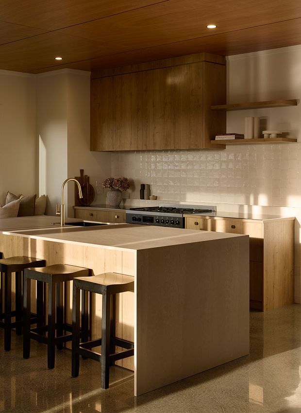 Warm contemporary kitchen with a large pale timber island and integrated seating, dark timber overhead cabinetry, open shelving, a brass-toned gooseneck faucet, stainless steel range, and glossy cream textured tile splashback. The space has a timber-lined ceiling, polished reflective flooring, recessed lighting, and a partially visible adjacent living area.