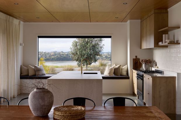 Warm, refined open-plan kitchen and dining space with a large central island, integrated window seat, timber cabinetry, white textured tile splashback, and a timber-lined ceiling. The wide picture window overlooks a calm waterfront landscape, while a dark timber dining table and sculptural ceramic vase occupy the foreground.