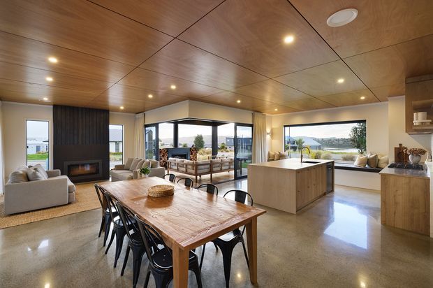 Open-plan contemporary living, dining and kitchen interior with a large timber dining table, black dining chairs, polished concrete flooring, timber-lined ceiling, central kitchen island and extensive glazed openings overlooking a landscaped exterior. A lounge area with fireplace is visible at left, while warm integrated lighting and natural timber create a refined, inviting atmosphere.