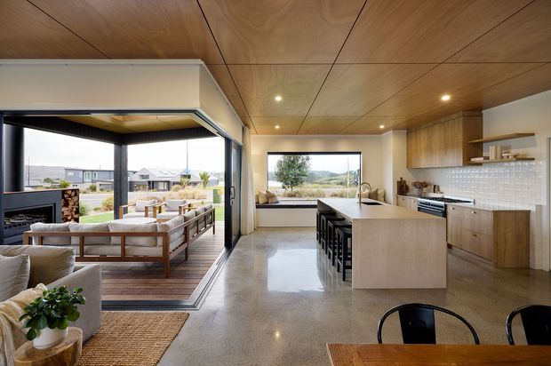 Open-plan contemporary kitchen, dining and living area with polished concrete flooring, extensive timber ceilings, and large glazed openings connecting to a covered outdoor terrace. The kitchen features a large pale timber island with black stools, timber cabinetry, a white textured tile splashback, and integrated appliances. A timber dining table sits in the foreground, while the adjacent living area includes a sofa and low timber-framed seating. The room has abundant natural light and views toward a suburban landscape.