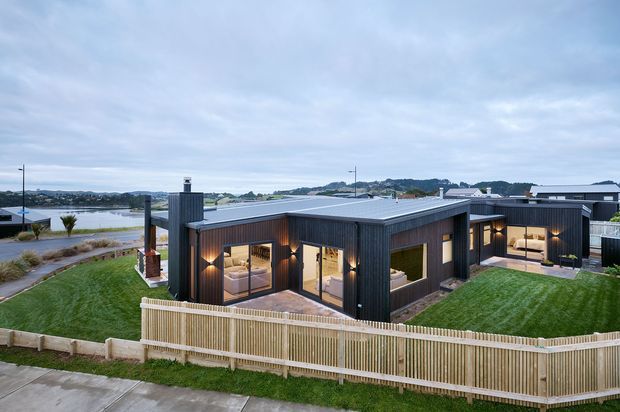 A contemporary single-storey dark-clad house viewed from above, with a low flat roof, extensive floor-to-ceiling glazing, warm interior lighting, paved outdoor areas, lawns, and a pale timber boundary fence. A waterfront or coastal inlet and neighbouring homes are visible in the background.