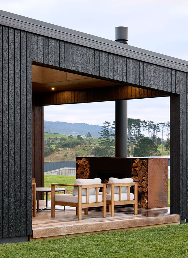 Exterior view of a contemporary dark-clad pavilion or house opening onto a covered outdoor living area. The sheltered space has timber decking, two light upholstered armchairs, a large built-in fireplace with stacked firewood and a cylindrical flue, and expansive views toward rolling countryside and trees. The building features vertically textured dark cladding, a simple low-pitched roof, and warm timber soffit detailing.