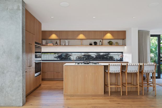 Contemporary open-plan kitchen with warm timber cabinetry, a large central island, integrated appliances, open display shelving, and a long horizontal splashback window. The island incorporates seating for four with woven-back timber bar stools. Concrete-textured walling and timber flooring add natural material contrast, while glazing at right brings in greenery and daylight.