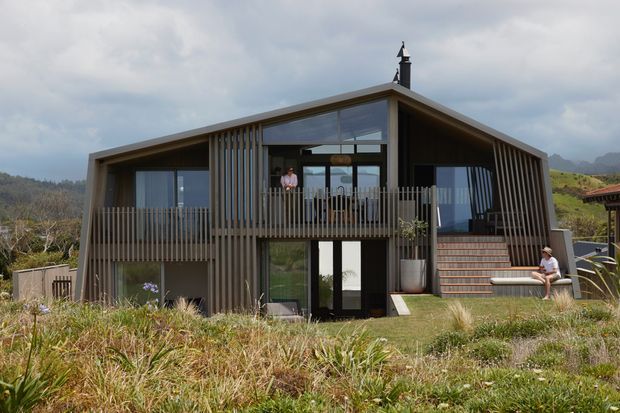 A contemporary two-storey timber-clad house set in a sloping natural landscape. The gabled roof has broad overhangs and a prominent chimney, while extensive glazing, vertical timber screening, and a raised balcony define the facade. A central glazed entry sits beneath the balcony, with exterior stairs leading to the upper level. Native grasses and planting surround the house, and distant hills are visible beyond.
