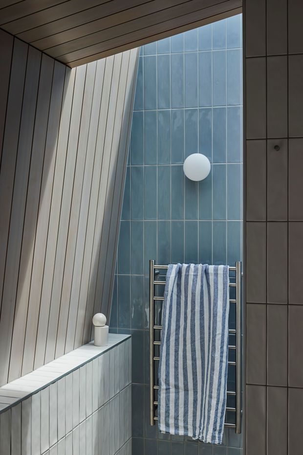 A compact, architecturally detailed bathroom interior featuring a tall blue glazed-tile wall, dark grey tiled side wall, pale vertically panelled wall and ceiling, and a stainless-steel heated towel rail with a blue-and-white striped towel. A small wall light and spherical lamp add understated detail. The space has a narrow, vertically emphasised composition with cool, muted tones.