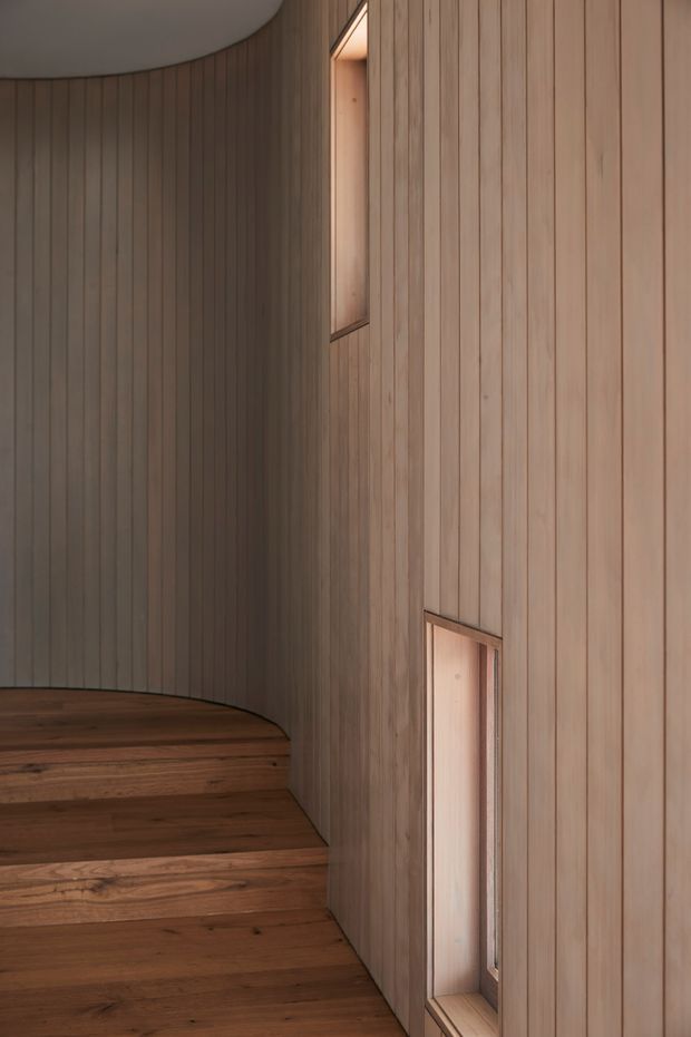 A close-up interior view of a curved stairway and adjoining wall, both finished in narrow vertical timber battens. Warm timber treads follow the rounded wall, while two narrow recessed openings provide subtle illumination. The space has a minimalist, refined and warm character with a smooth curved ceiling edge.