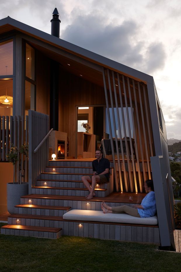 Exterior architectural photograph of a compact, multi-level contemporary house at dusk. The steeply sloped facade combines dark grey cladding, warm timber soffits and wall lining, extensive glazing, and a tall vertical timber privacy screen. Broad illuminated steps lead to a recessed entry and sheltered outdoor seating area, with a chimney visible above the roofline and distant hills beyond.