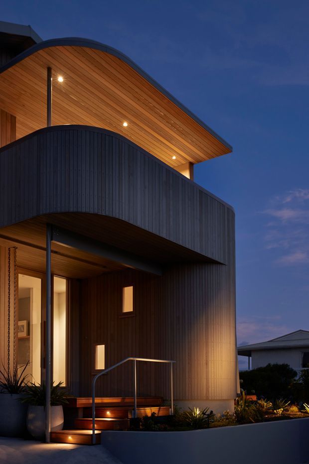 Twilight exterior view of a contemporary two-storey residence with a strongly curved roof canopy and matching curved upper facade. The building is clad in dark vertical boards, with a warm timber-lined soffit, recessed downlights, narrow windows, an illuminated entry, metal handrail, landscaped planter and steps. A neighbouring house is visible in the background.