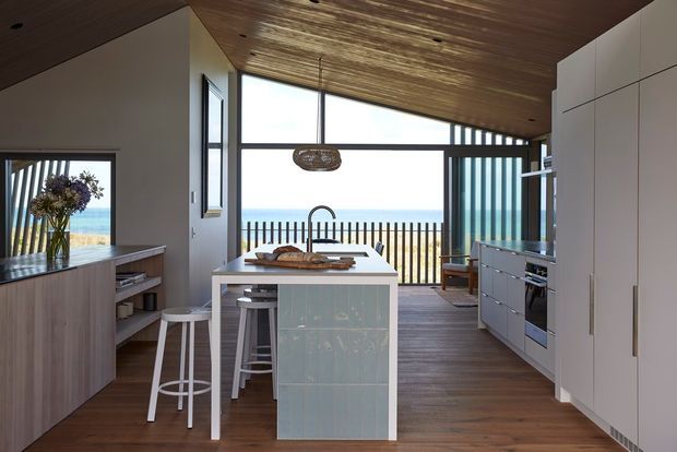 A bright contemporary coastal kitchen with a central island, integrated white cabinetry, timber flooring, and a slatted timber ceiling. Floor-to-ceiling glazing opens to a balcony or deck overlooking the ocean, while black-framed windows and doors create a strong architectural rhythm. The island includes seating, and a woven pendant adds a natural decorative touch.
