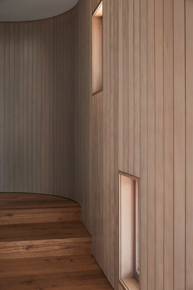 A close-up architectural interior view of a curved stairway with broad dark timber treads and vertically slatted timber wall panelling. Narrow recessed openings or windows punctuate the wall, creating warm, understated illumination. The palette is natural wood in warm brown and muted beige tones, with a smooth white curved ceiling edge visible above.
