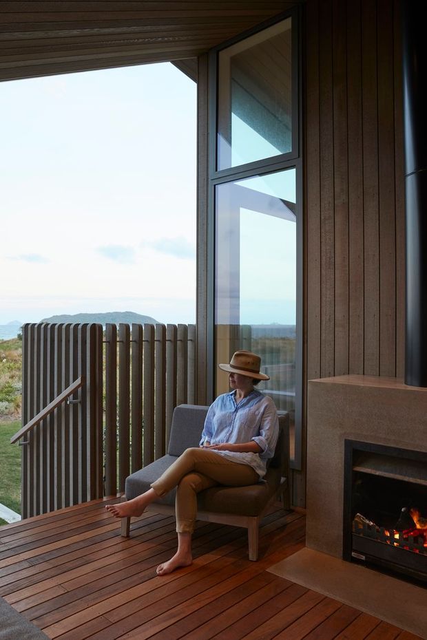 A warm, intimate indoor-outdoor living space with a person seated in an armchair beside a built-in fireplace. The room opens onto a timber deck and balcony with vertical timber screening, while large glazed doors and windows frame a distant natural landscape. Timber-lined ceilings and walls, dark window frames, and a simple earthy palette create a refined cabin-like atmosphere.