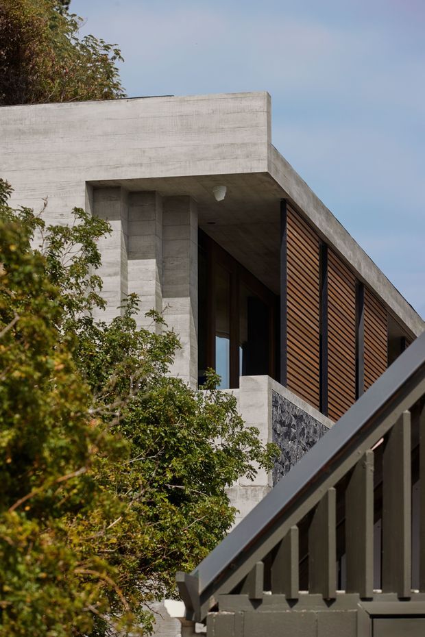 Exterior architectural view of a modern concrete residence, showing a flat roof, strong cantilevered geometric forms, deep recessed glazing, warm timber horizontal screening, and a dark stone accent wall. Vegetation partially frames the building, while a dark foreground railing crosses the lower right.