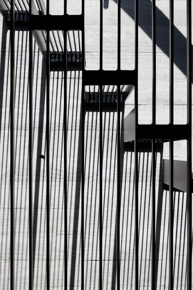 Abstract architectural detail showing strong sunlight casting long, repeating shadows from a black metal railing or screen onto a pale textured concrete wall and floor. The composition is highly geometric, with vertical bars, rectangular openings, and sharp black-and-white contrast; only partial architectural surfaces are visible, with no identifiable room or broader building context.
