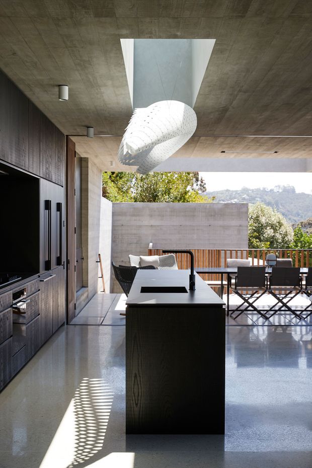 A modern open-plan kitchen and living area with a dark wood built-in media wall, central black kitchen island, polished concrete floor and ceiling, and expansive outdoor opening. A sculptural white pendant hangs beneath a large rectangular skylight. The space opens to a timber-railed terrace furnished with outdoor dining chairs, with trees and hills visible beyond.