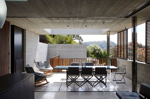 A modern, partially enclosed outdoor dining terrace or covered patio with a long dining table and dark folding chairs, lounge rocking chairs, and a built-in dark cabinet. Raw concrete walls and ceiling, timber screens, slender structural posts, and large openings frame a leafy mountainous view. The space has strong indoor-outdoor connection and abundant natural light.