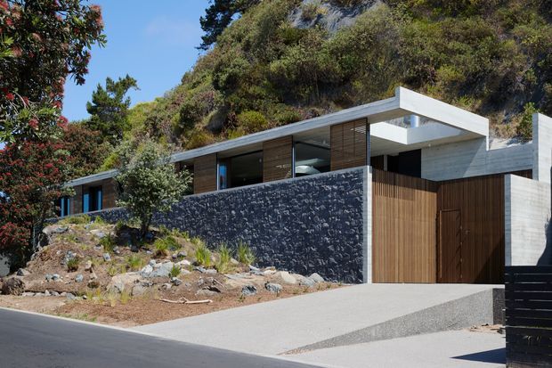 Contemporary hillside residence integrated into a steep, vegetated site. The low-profile building combines a dark rough-stone retaining wall, pale concrete roof and facade elements, extensive glazing, timber screening, and a tall timber entry gate. A sloped driveway and landscaped rocky foreground lead to the entrance, while dense native-looking vegetation rises behind the house.