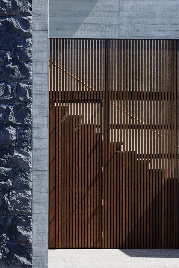 Close-up exterior view of a contemporary building facade combining rugged dark stone masonry, pale board-formed concrete, and a tall timber-look vertical slatted screen. The slatted screen conceals a glazed or recessed entry and reveals an internal stairway behind it. Strong sunlight creates geometric shadows across the facade and screen.