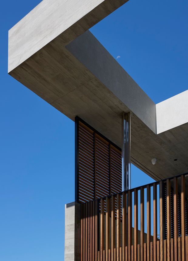Upward-looking exterior architectural photograph of a contemporary concrete building with strong cantilevered roof planes, deep concrete soffits, dark timber louvres, and a vertical timber slat screen against a clear blue sky.