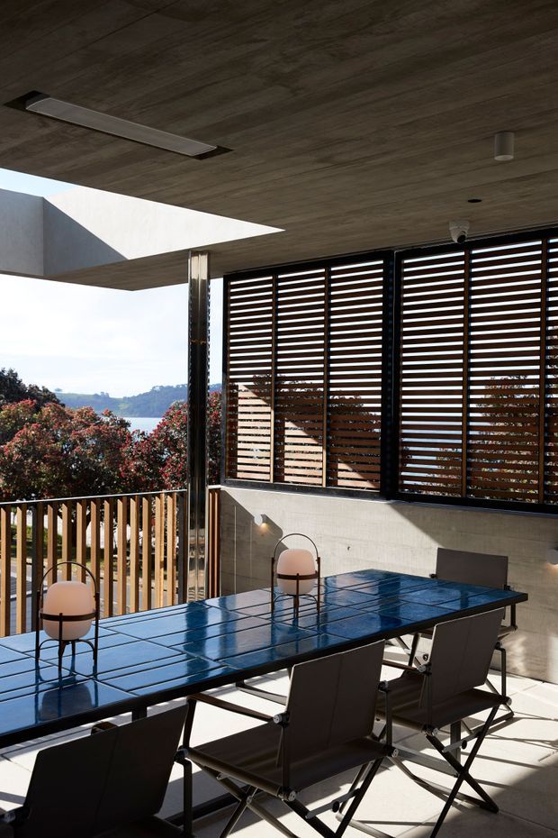 A modern, partially enclosed outdoor dining terrace with a long glossy blue tiled dining table and dark contemporary chairs. Timber horizontal louvres screen the glazing, while a timber-lined flat soffit, concrete walls, slim metal framing, and a timber balustrade define the space. The terrace overlooks a leafy, mountainous landscape and is lit by strong natural daylight.