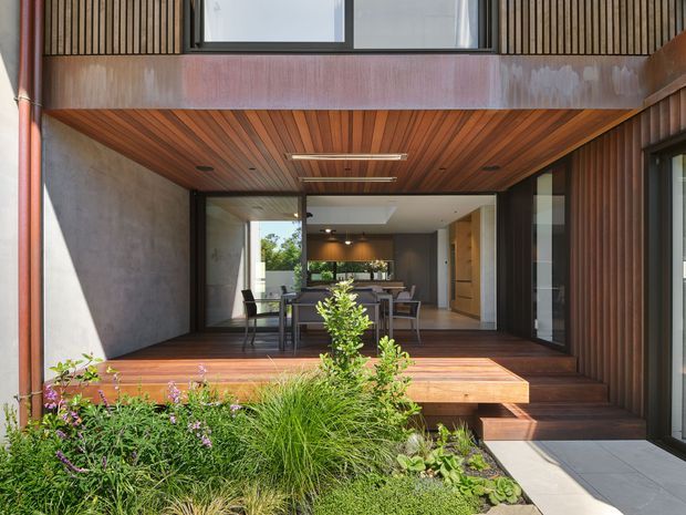 A contemporary residential courtyard or outdoor entertaining area framed by a timber-clad house. Large glazed sliding doors open onto a raised timber deck with outdoor dining furniture, while the covered soffit and deck ceiling feature warm timber lining and integrated linear lighting. Vertical timber battens, dark window frames, concrete-look wall surfaces, and lush planted landscaping create a warm indoor-outdoor connection. The interior kitchen and dining area is visible through the open glazing.