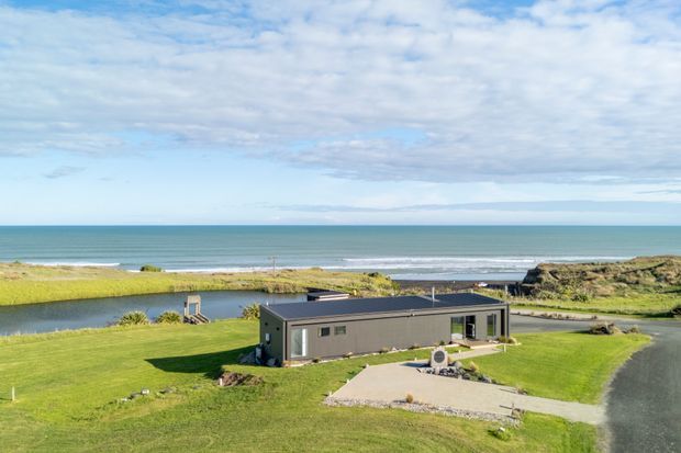 Aerial exterior view of a low, elongated modern building set in a broad grassy coastal landscape. The dark grey rectangular structure has a shallow flat roof, large windows, and a paved forecourt, with a lagoon or inlet and ocean visible directly beyond it.