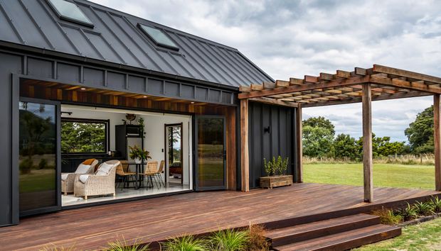 A modern single-storey dwelling with dark standing-seam metal cladding and a steeply pitched roof incorporating skylights. Large glazed sliding doors open from the interior onto a broad timber deck, which continues beneath a timber pergola. The deck overlooks an expansive grassy landscape, while the interior reveals a light-filled living and dining area with timber ceiling lining and warm furnishings.