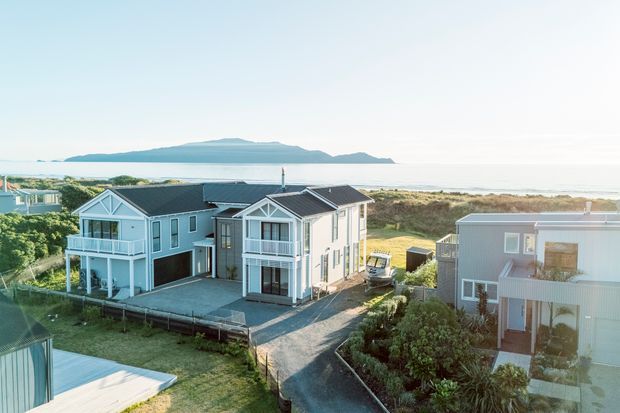 Aerial exterior view of a coastal residential development with several light-coloured multi-storey houses, dark pitched roofs, balconies, decks, and large glazed openings. The homes sit beside sandy coastal vegetation with ocean water and a distant island or headland visible beyond.