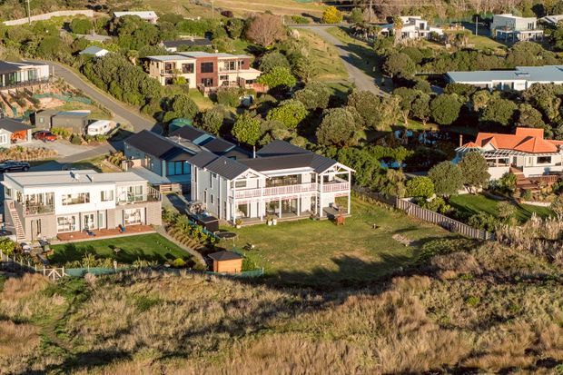 Aerial view of a suburban coastal residential neighbourhood with several detached homes set among mature trees, landscaped gardens, winding roads and open grassy lots. The central property is a large light-coloured multi-level home with dark pitched roofs, broad verandas and prominent white structural detailing. Surrounding homes show a mix of contemporary and traditional forms. Dune-like grasses occupy the foreground, while rolling terrain and distant vegetation extend behind the houses.