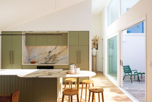 Bright contemporary kitchen with muted sage-green full-height cabinetry, a dramatic veined stone splashback, and a rounded marble-look island with timber stools. The kitchen opens to a tall glazed living or outdoor connection, with abundant natural light and a small deck visible beyond the sliding doors.