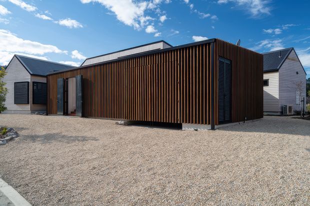 Exterior view of a contemporary single-storey building with a central rectangular volume clad in closely spaced vertical timber battens, dark louvred openings, and a low flat roof. Smaller gabled wings with light weatherboard cladding and dark pitched roofs sit on either side. The building fronts a broad gravel forecourt under a bright blue sky.