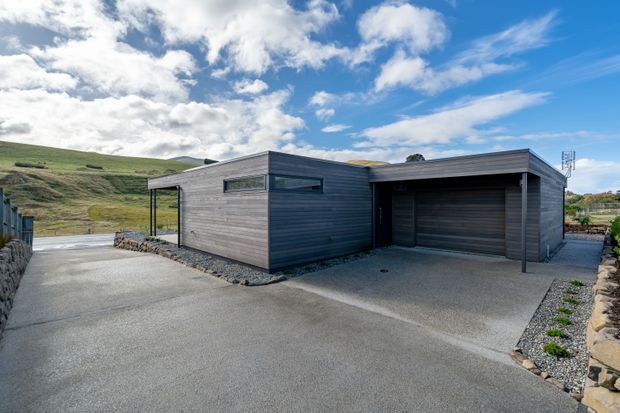 A low, angular contemporary house or ancillary building set in an open rural landscape. The exterior is clad in horizontal dark grey timber or composite boards, with narrow horizontal windows, a recessed glazed section, a flat or very low-pitched roof, and an integrated single garage with a dark sectional door. A broad concrete driveway and gravel-edged landscaping surround the building, with stone retaining walls and rolling grassy hills beyond.