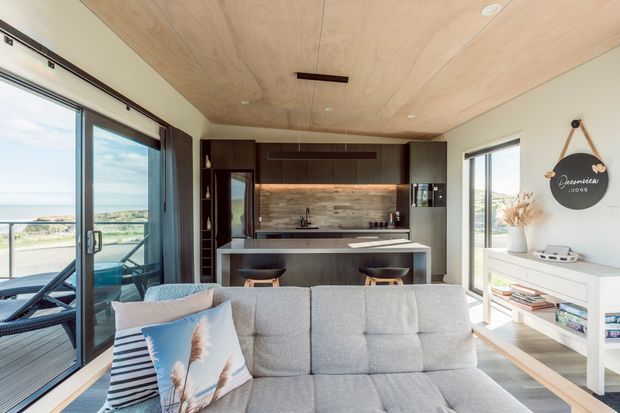 Bright open-plan living area with a compact contemporary kitchen and island behind a large light-grey sofa. The room has extensive glazing and sliding doors opening to a balcony with a coastal view, a light timber-lined ceiling, dark timber cabinetry, a stone-look splashback, and white built-in media/storage furniture. Coastal accents and abundant daylight create an airy, relaxed interior.
