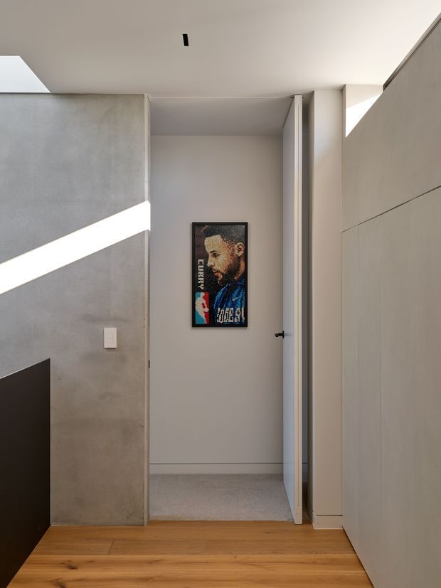 A minimalist interior view looking toward an open doorway and a small carpeted room or wardrobe area. The space features warm timber flooring, smooth light-grey plaster or concrete walls, flush pale cabinetry on the right, a simple white door, and strong angular sunlight entering from the left. A framed portrait artwork is mounted on the rear wall.
