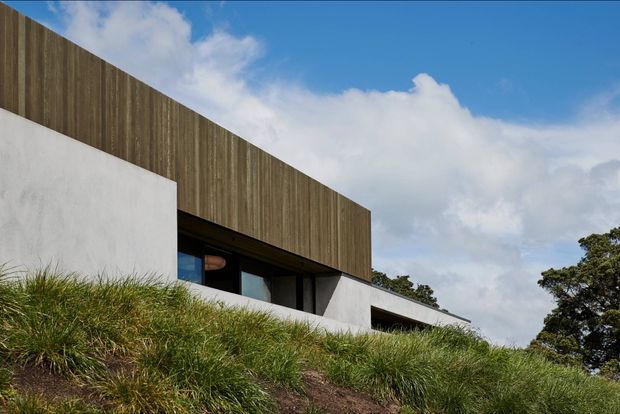Exterior view of a contemporary, low-profile building set into a grassy sloping landscape. The facade combines vertical timber cladding with smooth white rendered walls and dark recessed glazing beneath a projecting upper volume. Blue sky, clouds, and mature vegetation surround the structure.
