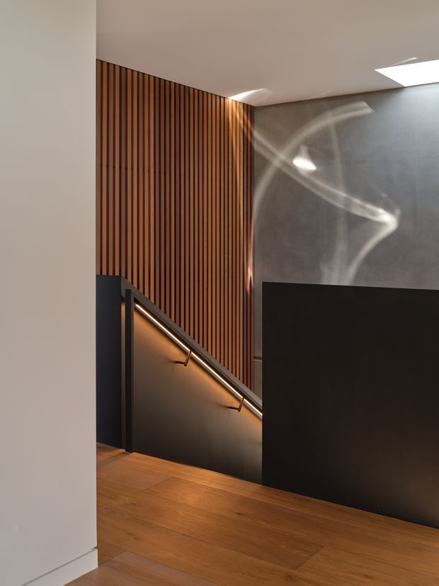 A refined modern stairway viewed from a hallway or doorway. Warm timber flooring continues toward a dark stair with slim metal handrails, while a vertical timber slat feature wall rises beside it. A textured grey wall and concealed/skylight-like ceiling openings create dramatic shafts of natural light. The palette is warm wood, charcoal, grey, and off-white, with integrated warm stair lighting.