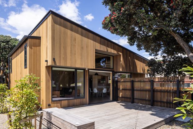 Exterior view of a contemporary timber-clad residence with a steep gabled roof and a high, recessed upper-level opening above glazed living and dining spaces. A broad timber deck, vertical slatted privacy fencing, and established landscaping frame the facade.