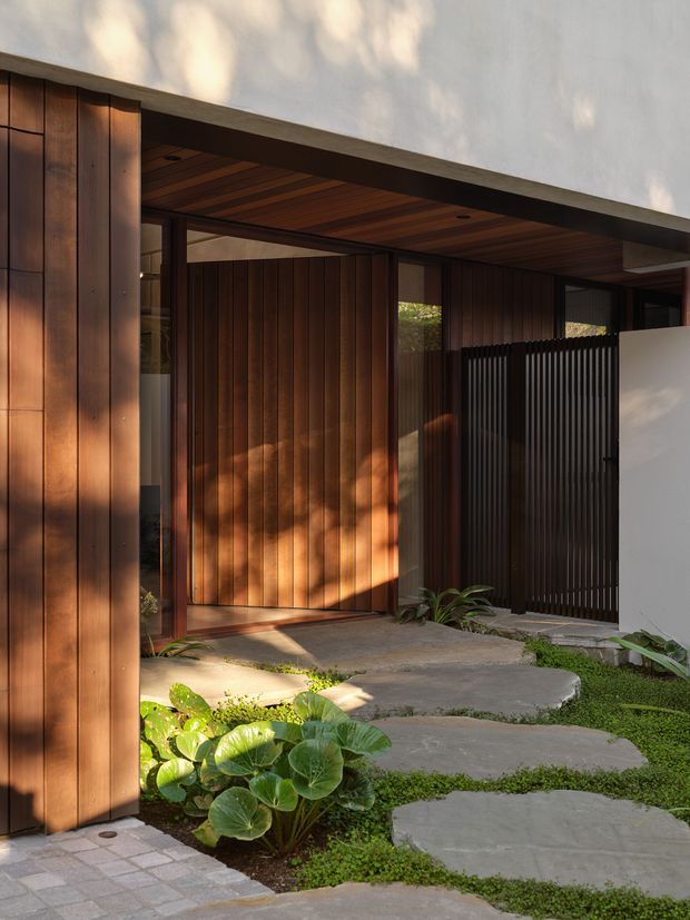 A contemporary residential entrance viewed from outside, featuring warm vertical timber wall cladding and a large timber pivot door beneath a deep timber-lined soffit. Dark vertical metal screening frames the entry, while pale rendered walls and irregular concrete stepping slabs lead through a lush, low planted landscape. The composition has strong indoor-outdoor character, warm sunlight, and a refined modern palette.