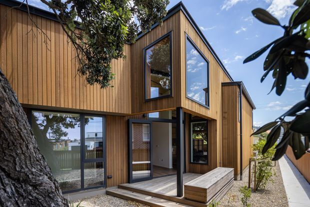 Exterior view of a contemporary two-storey timber-clad residence with pronounced box-like volumes, large dark-framed windows, a recessed entry, and a timber deck and built-in bench. The house sits on a narrow landscaped site with gravel paving and mature trees, under a bright blue sky.