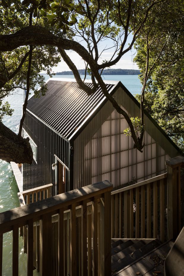 Exterior view of a compact waterside building nestled among mature trees, photographed from a timber stair or deck. The structure has a steep gabled roof clad in corrugated metal and dark vertical siding, with translucent or light-toned panel cladding on the gable end. A narrow timber jetty or deck borders the water, with a forested shoreline visible across the bay.