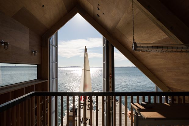 Interior view from an upper-level landing or loft within a compact timber-clad structure, looking through a large triangular gable opening toward sparkling waterfront. A sailboat is visible outside, with a timber deck and dark balustrade in the foreground. The interior has exposed plywood or timber surfaces, a steep pitched roof, exposed rafters, and simple utilitarian lighting and shelving.
