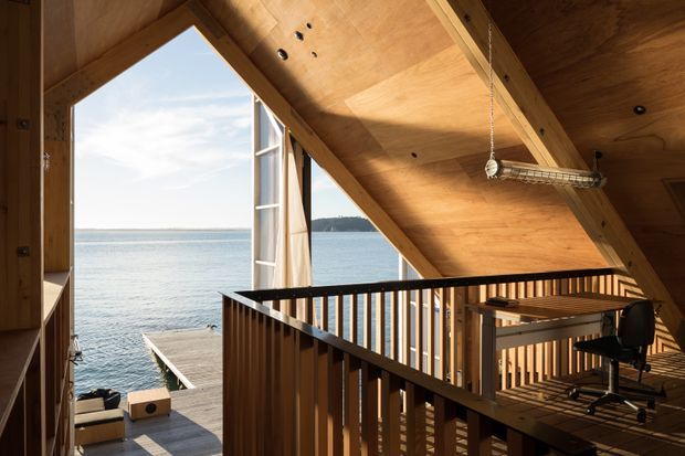 A warm timber-lined loft or upper-level interior overlooking calm coastal water through large angular glazing. The space features exposed timber roof framing and plywood-like ceiling surfaces, a black metal balustrade around a stair or mezzanine opening, and a compact built-in desk with an office chair. A deck and waterfront are visible beyond the open glazing.