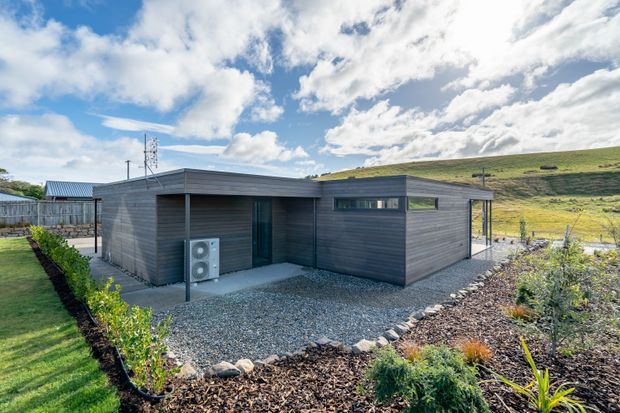 Single-storey contemporary low-profile house set in a landscaped, sloping rural site. The exterior is composed of dark horizontal timber-like cladding, flat roof planes, recessed glazing and a sheltered entry/courtyard zone. Gravel surrounds the building, with planted garden beds and a fenced grassy hillside in the background. Bright daylight and strong blue-sky contrast emphasize the clean, modern form.